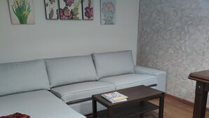 A living room with a grey sofa, a wooden coffee table, and a small table with a book. There are floral paintings on the wall.