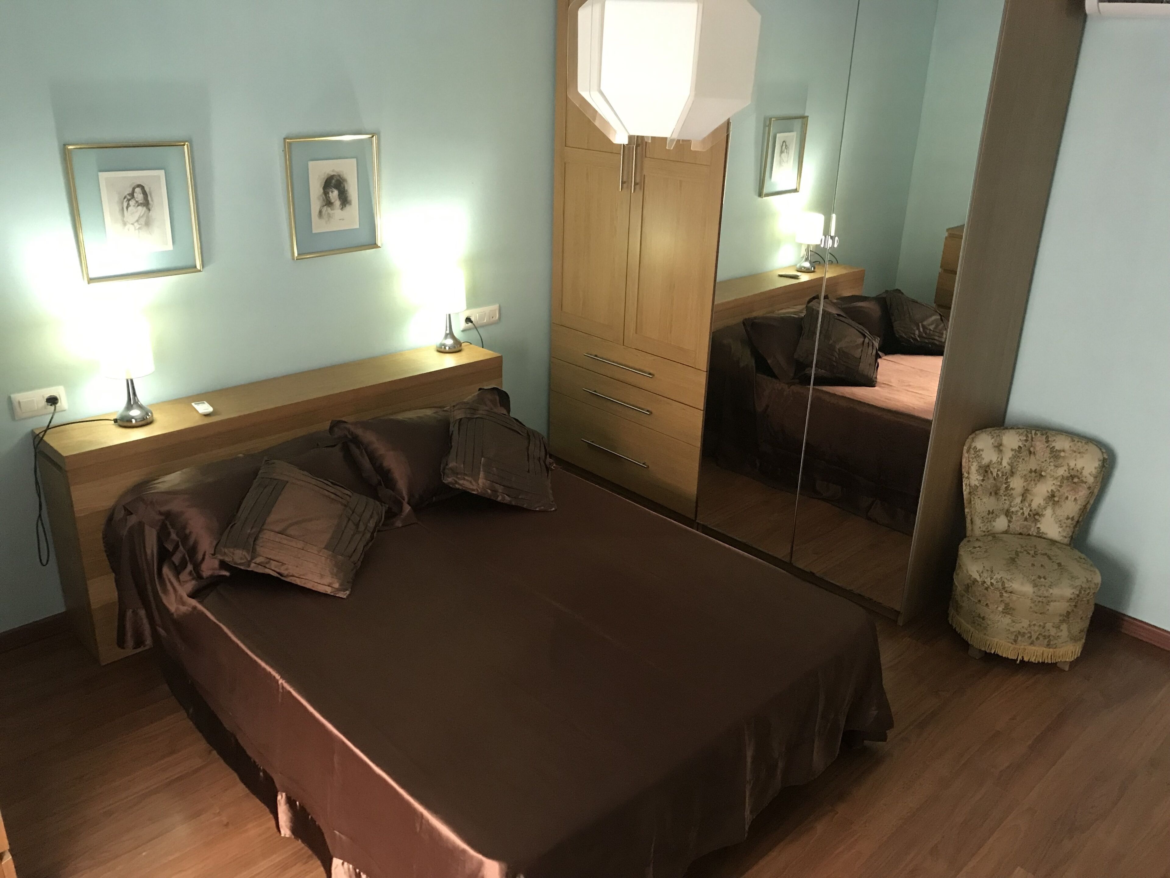 Un dormitorio con una cama, mesitas de noche, un espejo, una silla y dos cuadros en la pared.