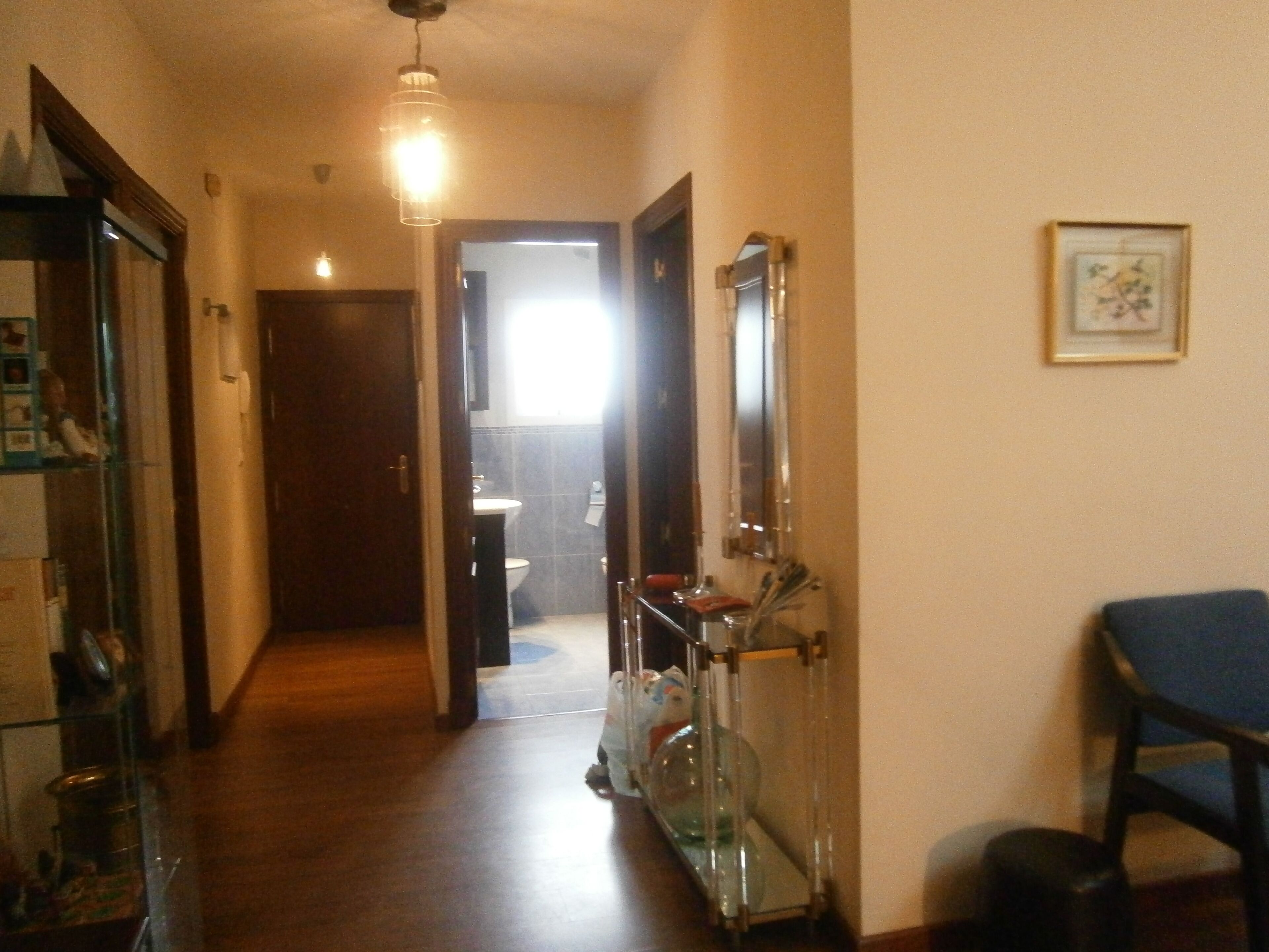 A hallway with a wooden floor, a glass display case, a mirror, and a blue chair.
