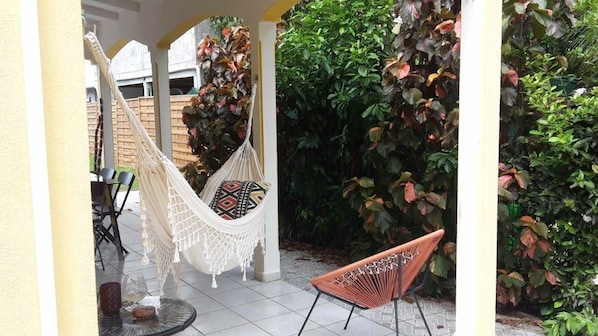 A hammock chair with a patterned cushion, a wicker chair, and a patio area with a tiled floor and a white wall.