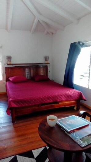 A bedroom with a wooden bed, a red bedspread, a small table with a cup and a book, and a window with curtains.