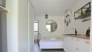 Een moderne, goed verlichte slaapkamer met een bed, een ronde spiegel en een kitchenette met witte kastjes en een houten aanrecht.