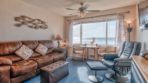 Ein gemütliches Wohnzimmer mit Ledersofa, Esstisch und Relaxsessel. Eine Deckenventilatoren und Blick aufs Meer sind durch die großen Fenster sichtbar.
