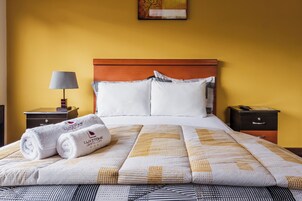 Una cama bien hecha con una colcha estampada, dos almohadas y dos toallas enrolladas sobre una mesita de noche.