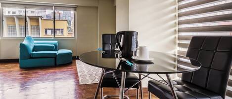 A modern living room with a round glass table, black leather chairs, a teal sofa, and a large window with blinds.
