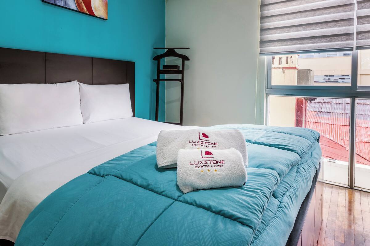 Una cama bien hecha con sábanas blancas y una colcha color turquesa, dos toallas con el logotipo 'LUXSTONE', un cabecero oscuro y una ventana con persianas.