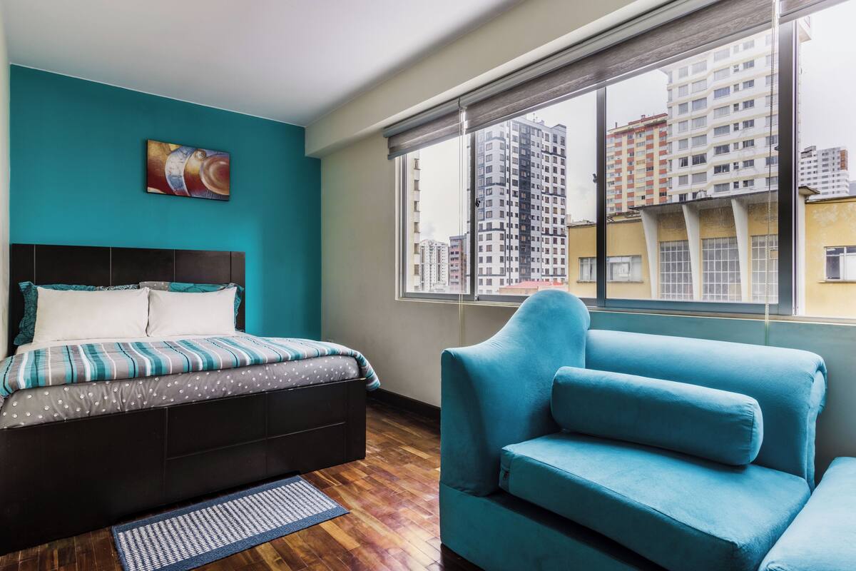 Un dormitorio con cama, un sillón azul, una ventana con vistas a la ciudad y un cuadro en la pared.
