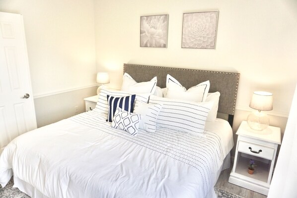 A neatly made bed with white bedding and striped pillows, a bedside table with a lamp, and two framed floral artworks on the wall.