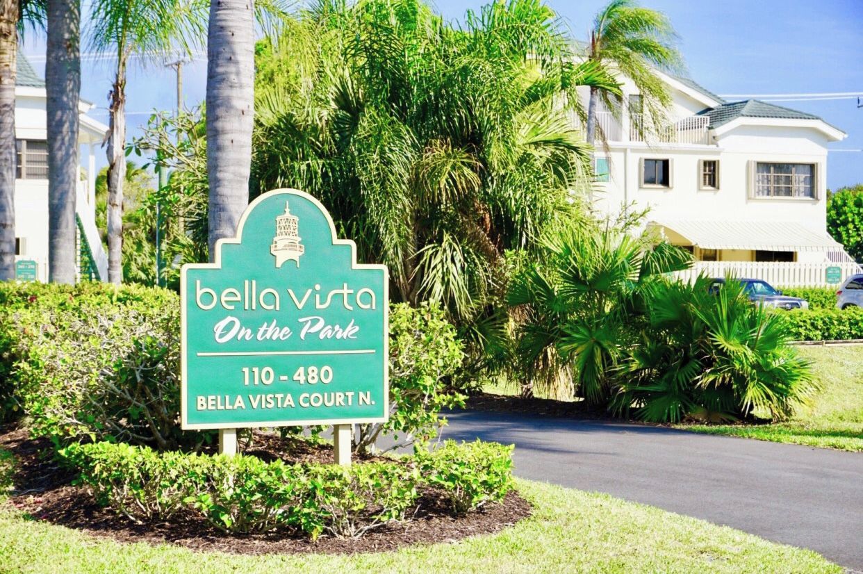 Un panneau indiquant « Bella Vista Court N. », avec un fond vert et des lettres blanches, entouré d’une végétation luxuriante et d’un bâtiment blanc en arrière-plan.