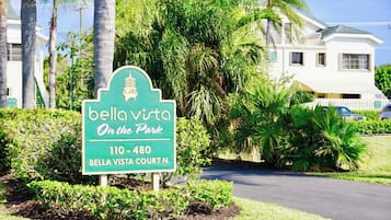 Un panneau indiquant « Bella Vista Court N. », avec un fond vert et des lettres blanches, entouré d’une végétation luxuriante et d’un bâtiment blanc en arrière-plan.