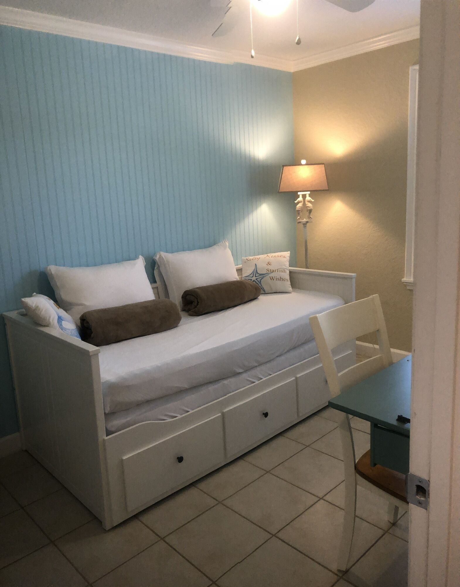 Habitación individual con una cama blanca, dos almohadas, un edredón marrón, una mesita de noche con una lámpara y una pequeña mesa.