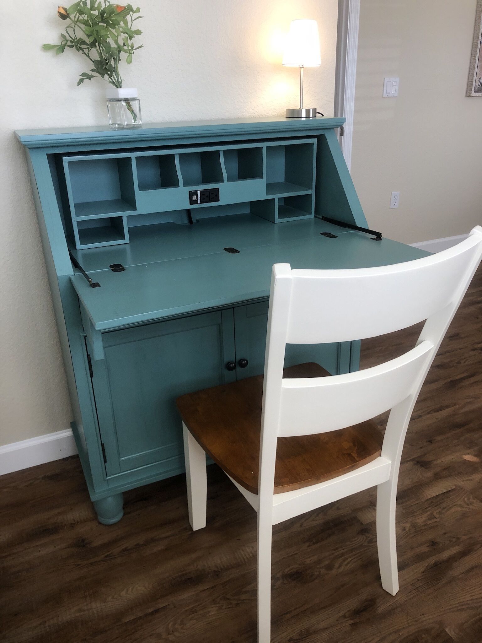 A teal desk with a white chair, a small vase with flowers, and a lamp on the desk.