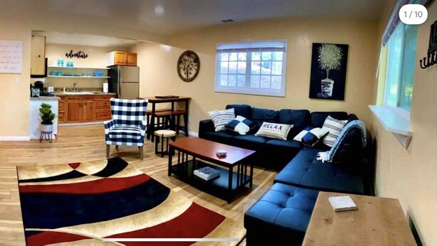 A living room with a sofa, coffee table, and a rug. There is a kitchen area with wooden cabinets and a refrigerator. - All New Remodeled Cottage (Three Rivers)