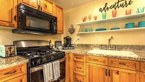 A kitchen with wooden cabinets, a black microwave, a stove, a sink, and a shelf with colorful glassware. - All New Remodeled Cottage (Three Rivers)