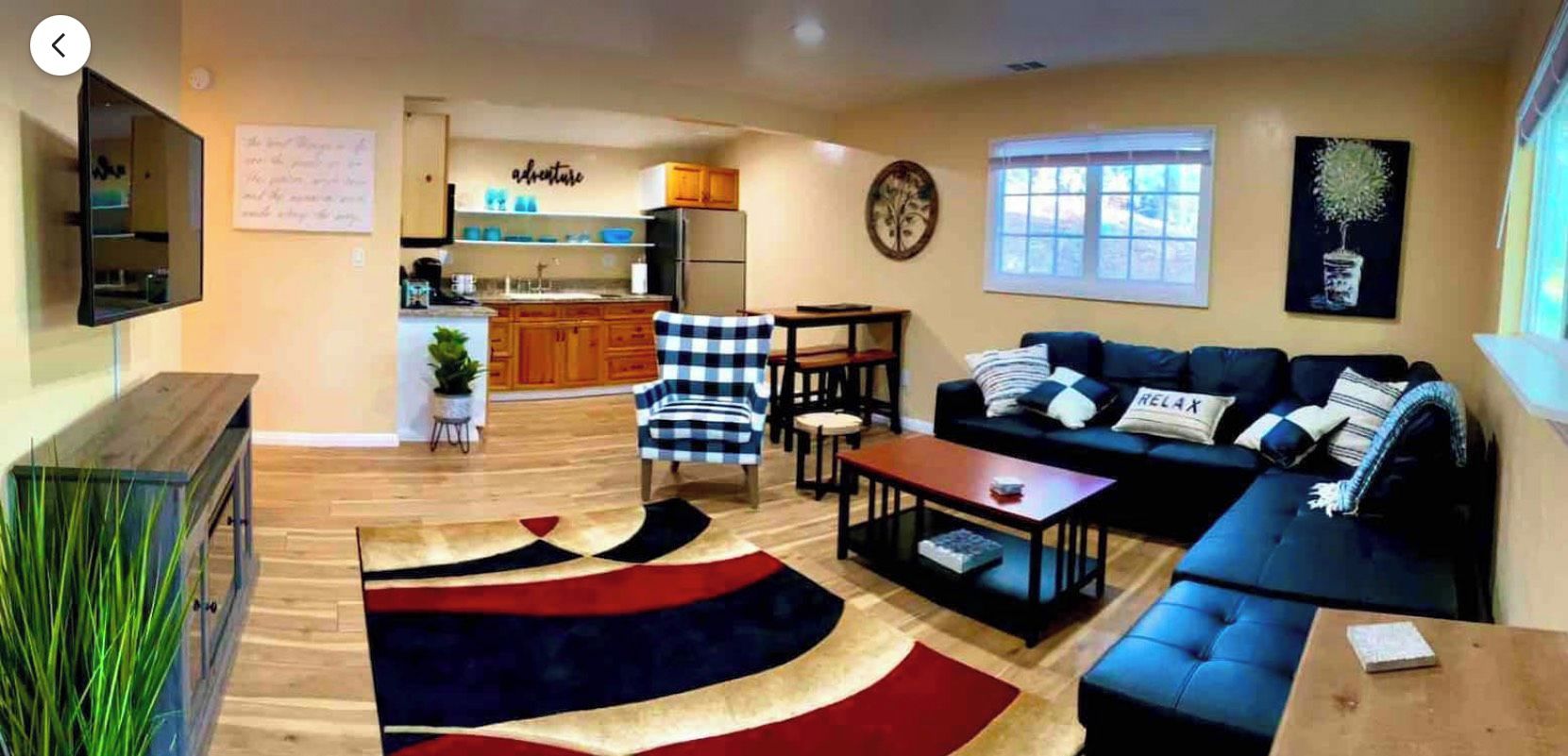 A living room with a sofa, coffee table, and a rug. There is a kitchen area with wooden cabinets and a refrigerator.