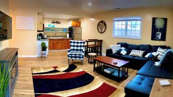 A living room with a sofa, coffee table, and a rug. There is a kitchen area with wooden cabinets and a refrigerator.