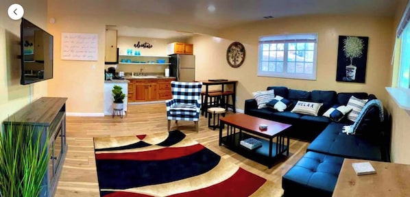 A living room with a sofa, coffee table, and a rug. There is a kitchen area with wooden cabinets and a refrigerator.