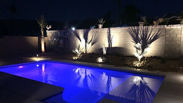 Una piscina con borde iluminado en azul, rodeada por un muro bajo con plantas en macetas y un muro decorativo.
