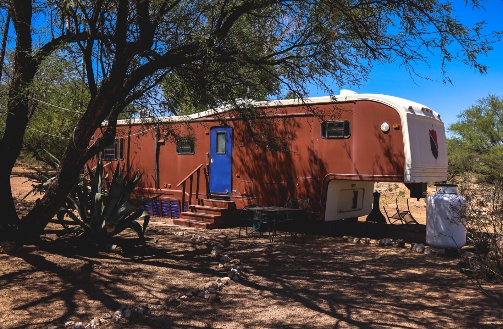 A stationary RV with a blue door, surrounded by trees and a clear sky.