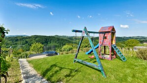 Un terrain de jeux équipé de balançoires et d’un toboggan, entouré d’une pelouse et offrant une vue sur des collines.