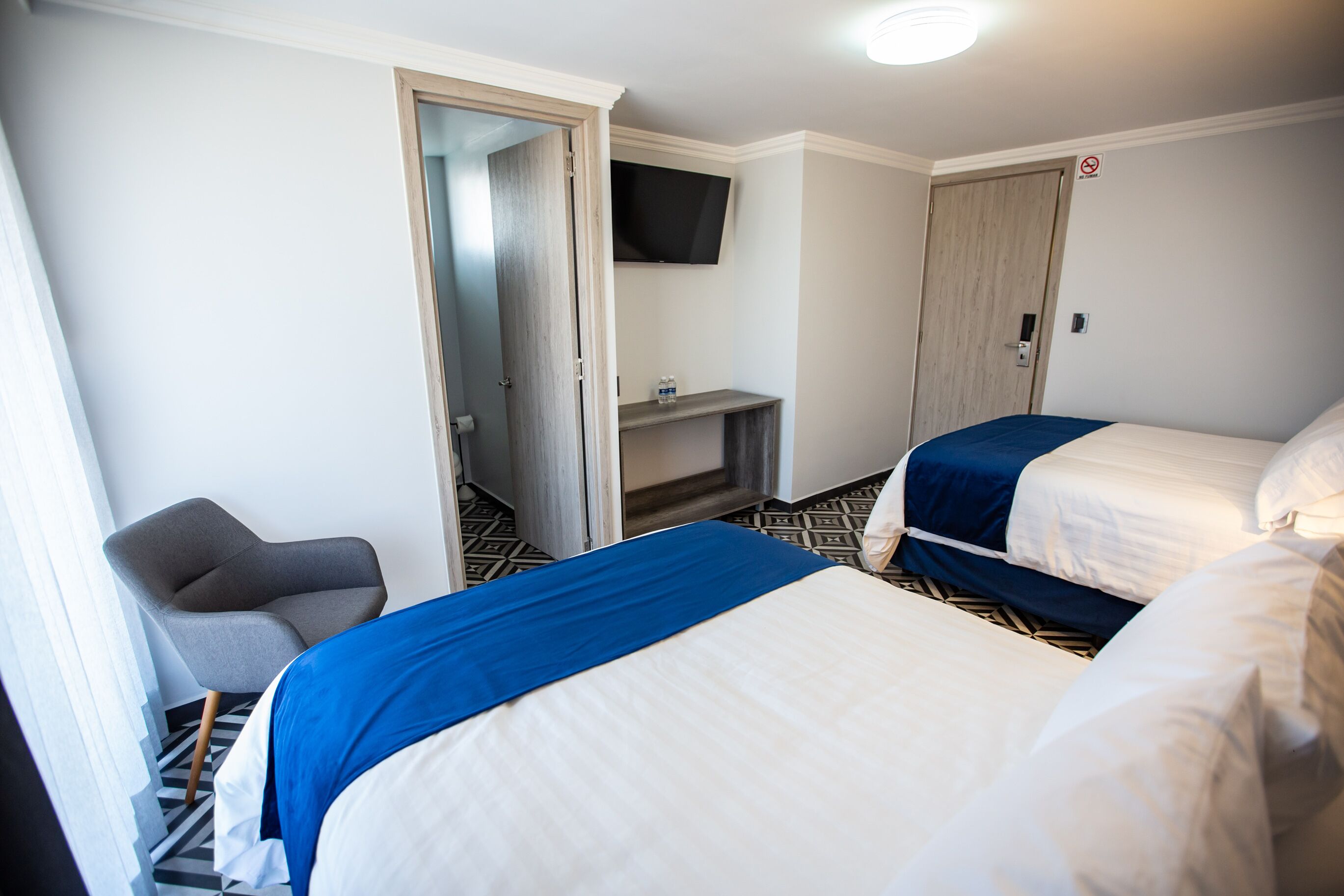 A hotel room with two beds, a TV mounted on the wall, a chair, and a door leading to another room.