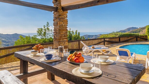 An outdoor dining area with a stone pillar, a wooden table set with bowls, glasses, and fruit, overlooking a pool and scenic hills. - Finca Hornillo Cómpeta by Ruralidays (Málaga)