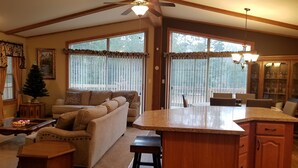 A spacious living room with a large sofa, a coffee table, and a small dining area with a table and chairs. There is a kitchen area with a countertop and cabinets. - New on market.  Cabin with 3 beds and 3 baths surrounded by forest (Grand Marsh)