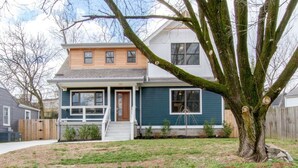 A two-story house with a front porch, a wooden fence, and a large tree in the yard.