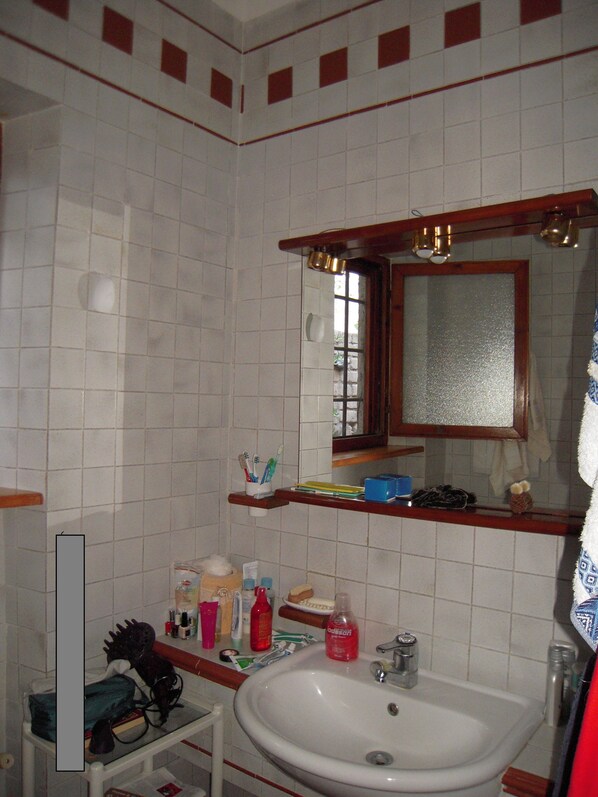 Une salle de bain avec un lavabo blanc, un miroir et des murs carrelés. On y trouve des étagères avec des articles de toilette et un porte-serviettes.