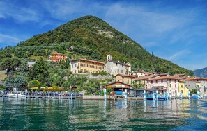 Un paese sul lago con edifici su una collina, barche ormeggiate lungo la riva e un cielo sereno.