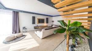 Un salon moderne avec un canapé, une table basse et un téléviseur fixé au mur. Un grand pot de plante se trouve dans un coin, et une petite table porte un panneau.