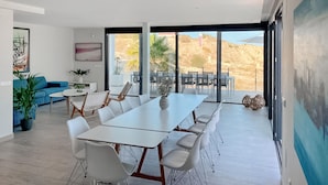 Una sala de estar moderna con una amplia mesa de comedor, sillas blancas y un sofá azul. Hay un ventanal con vistas a un paisaje desértico.