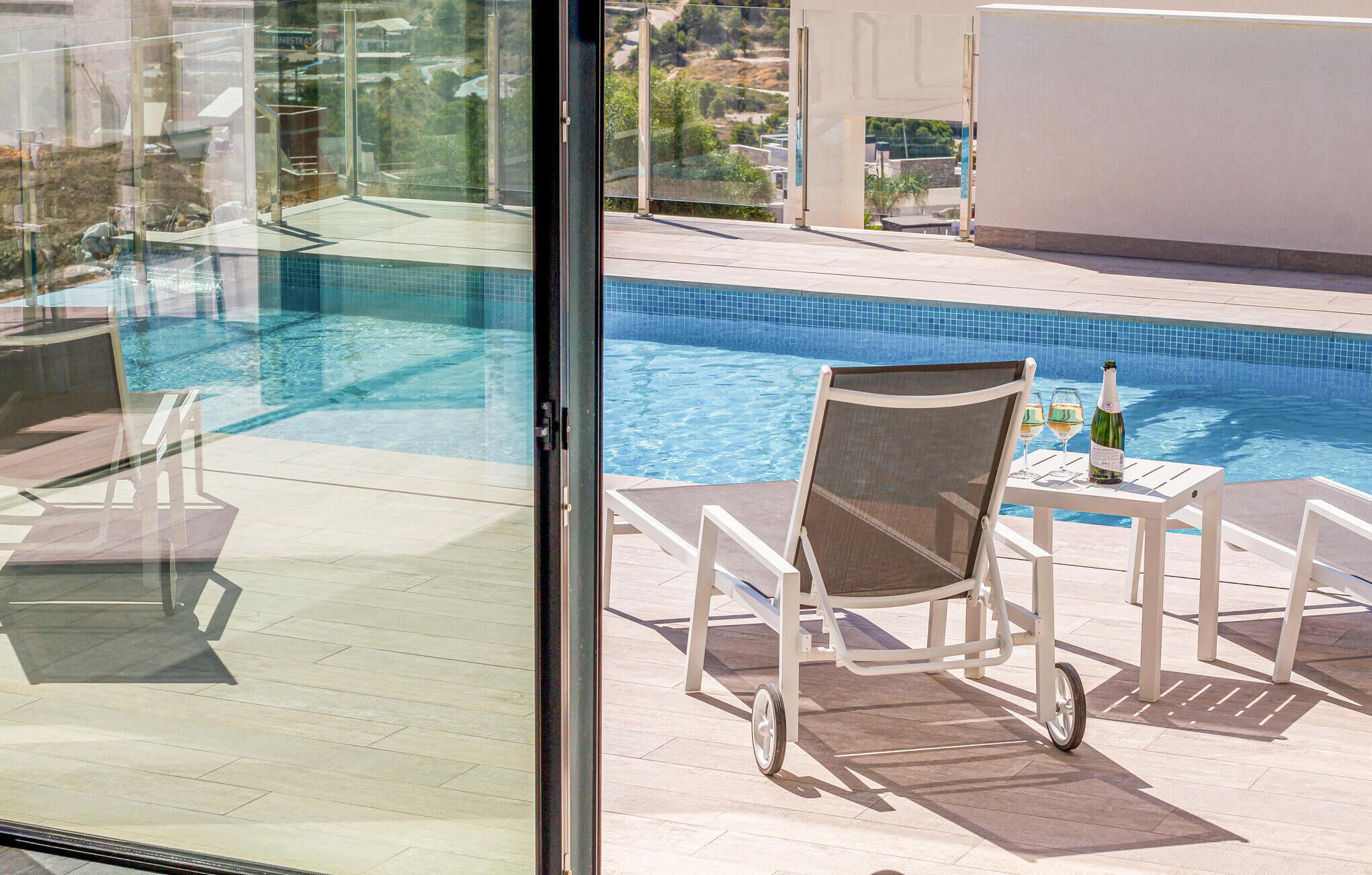 A poolside area with a glass door, a chair, a table with a bottle and glasses, and a view of a swimming pool.