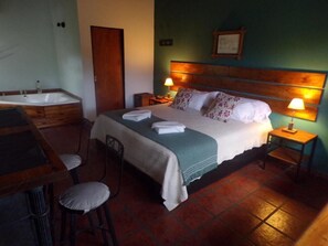 Un dormitorio con cama, lámparas de noche, cabecero de madera, una mesita, una silla y un baño con bañera.