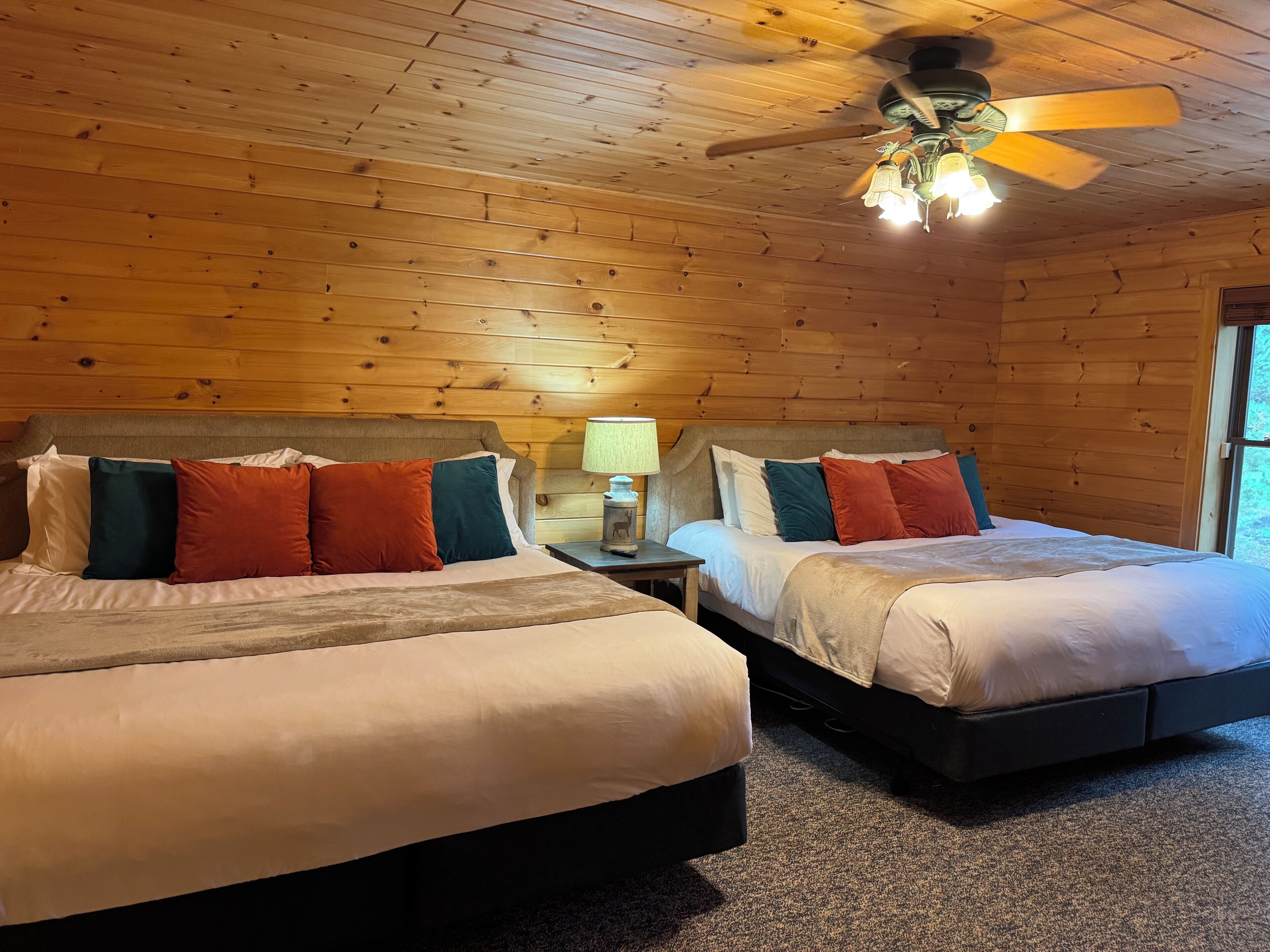 A room with two beds, wooden walls, a ceiling fan, and a nightstand with a lamp.
