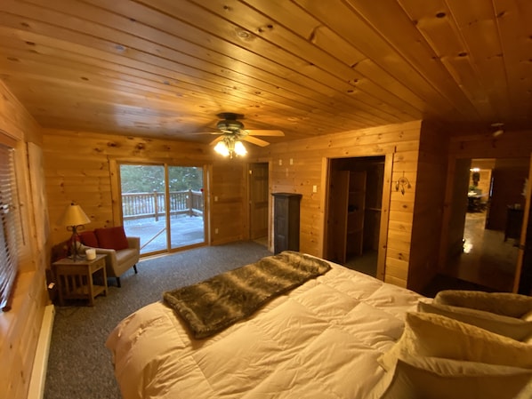 A wooden cabin room with a bed, a small table with a lamp, a fireplace, and a sliding glass door to a deck.