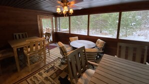 A wooden deck with a round table and chairs, a ceiling fan, and large windows offering a view of trees.