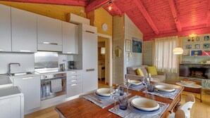 Une cuisine moderne avec des meubles blancs, une table à manger en bois pour quatre personnes et un coin salon avec un canapé et un téléviseur.