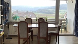Un coin repas comprenant une table et des chaises, un radiateur, une lampe sur pied et une vue sur un paysage rural grâce à de grandes fenêtres.