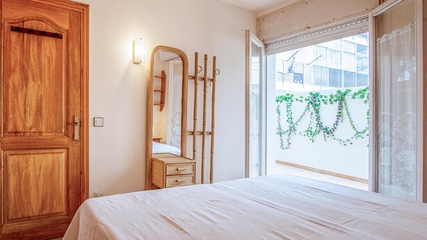 A bedroom with a wooden door, a mirror, a bed with a white bedspread, and a window with a view of greenery outside. - Beach front apartment in Sant Antoni de Calonge (Sant Antoni de Calonge)