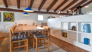 Un coin repas rustique avec une table et des chaises en bois, une suspension bleue et un mur blanc orné d’étagères où sont rangés des ustensiles de cuisine.