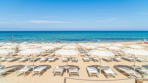 Ein Strand mit Reihen von Liegen und Sonnenschirmen, einem wolkenlosen, blauen Himmel und dem Meer im Hintergrund.