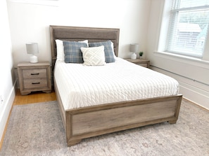 A bedroom with a wooden bed, two bedside tables, a window with blinds, and a rug. - Loft on Lincoln Hancock #204 (Hancock)