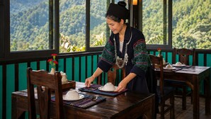 A person setting a table with traditional dishes and a view of a mountainous landscape through large windows. - Pu Hmong Eco Retreat (Sa Pa)
