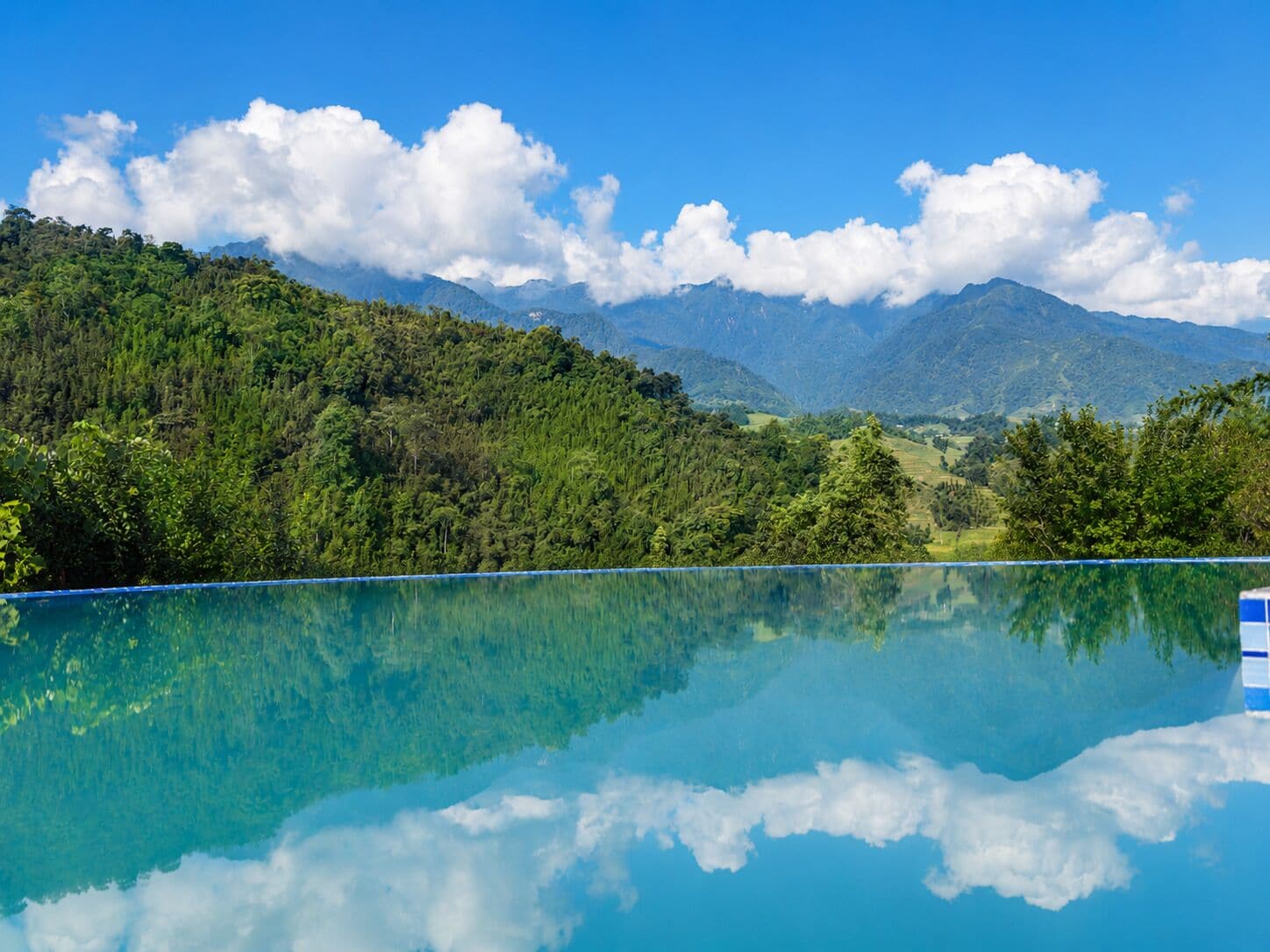 A clear blue pool reflecting the surrounding landscape, including lush greenery and distant mountains.