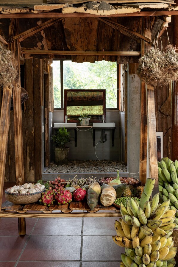 Une structure rustique en bois, avec un coin cuisine visible à travers une fenêtre, entourée de divers fruits et légumes disposés sur une table en bois.