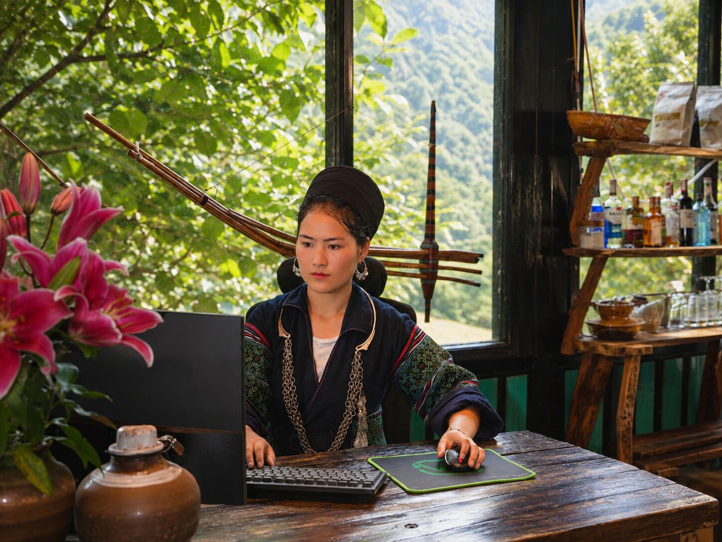 Uma pessoa sentada à mesa de madeira com um laptop, cercada por decoração tradicional e vista para o verde.