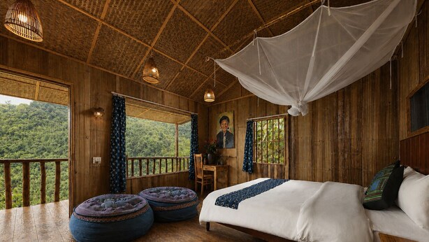 A room with a bed, two ottomans, a desk, and a chair. There is a large window with a view of greenery outside. - Pu Hmong Eco Retreat (Sa Pa)