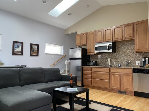 A modern kitchen with wooden cabinets, a stainless steel refrigerator, and a microwave. There is a gray sofa and a small table with a bottle on it.