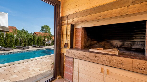 Ett utomhuskök i trä med en inbyggd grill, ett område vid poolen med solstolar och en utsikt över ett hus i bakgrunden.